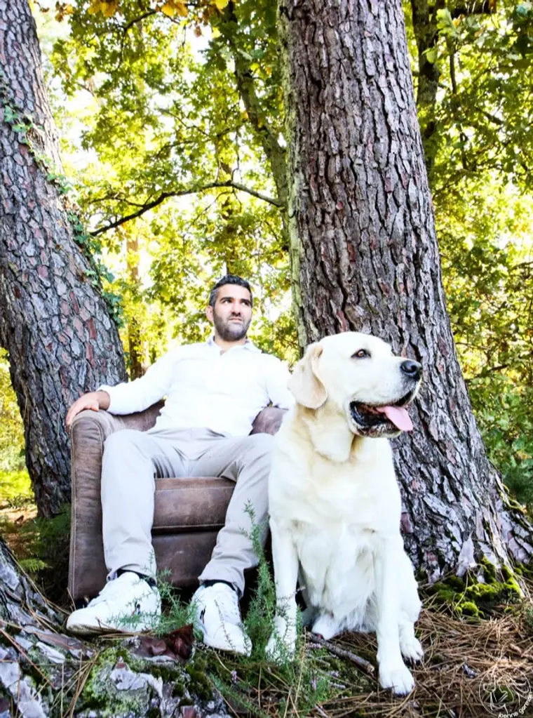 Homme assis dans un fauteuil au pied d'un arbre en forêt, son chien à ses côtés.