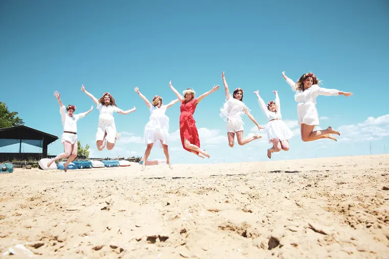 Groupe d'amies sautant de joie sur la plage, couronnes de fleurs, ciel bleu.