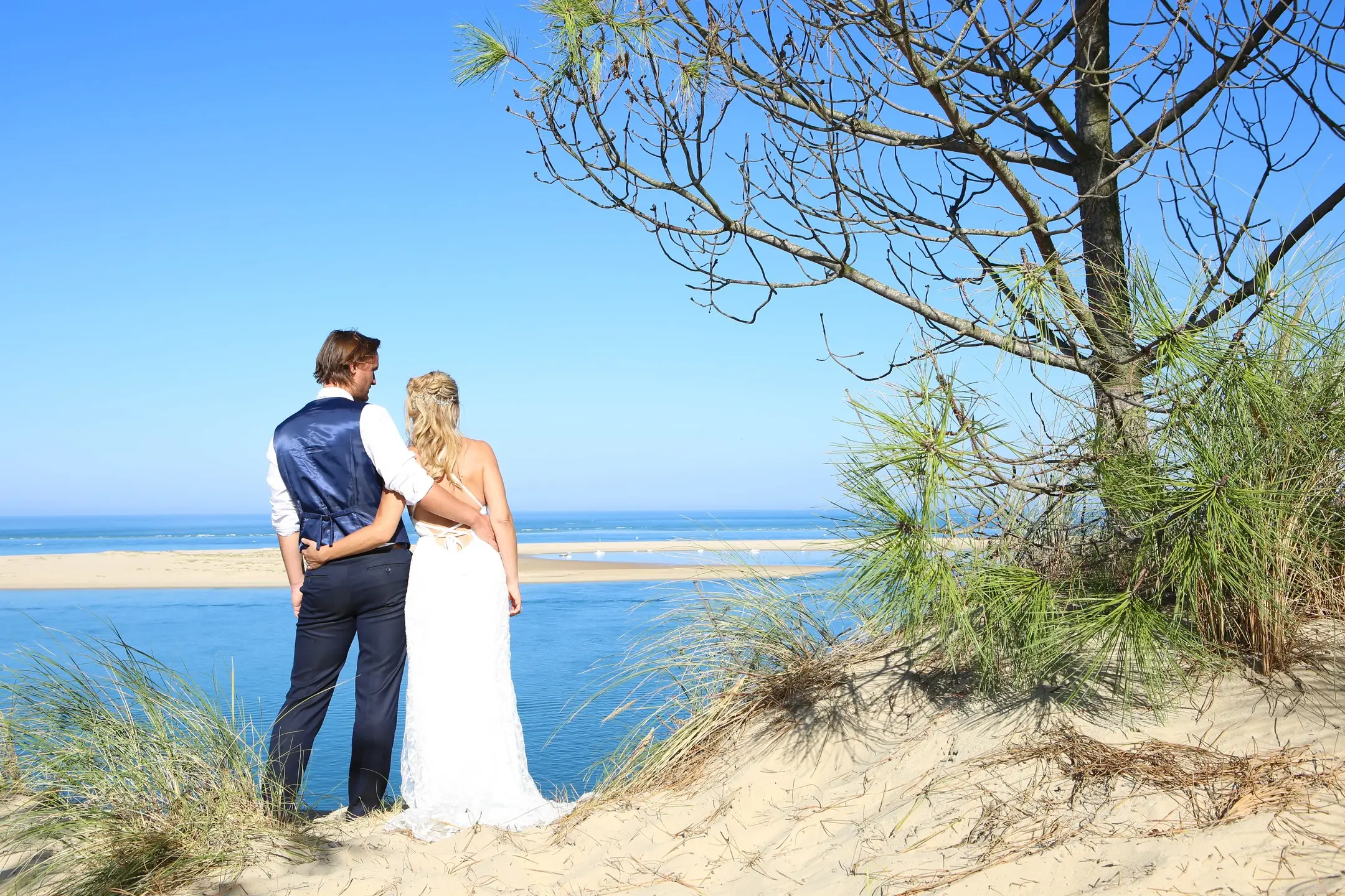 Un couple, de dos, contemple le Bassin d'Arcachon depuis une dune bordée de pins.